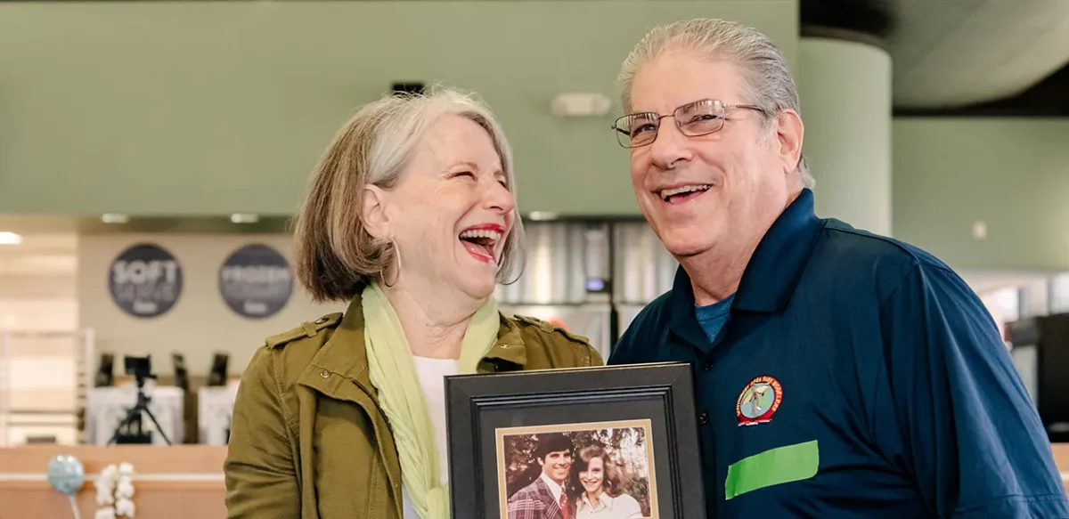 A man and a woman smile and laugh at one another while holding an aged photograph of themselves.