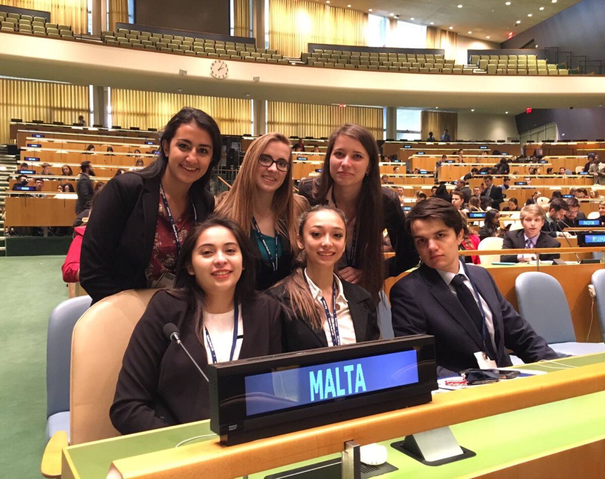 Six students sitting together in professional clothing in front of a sign designating "Malta"