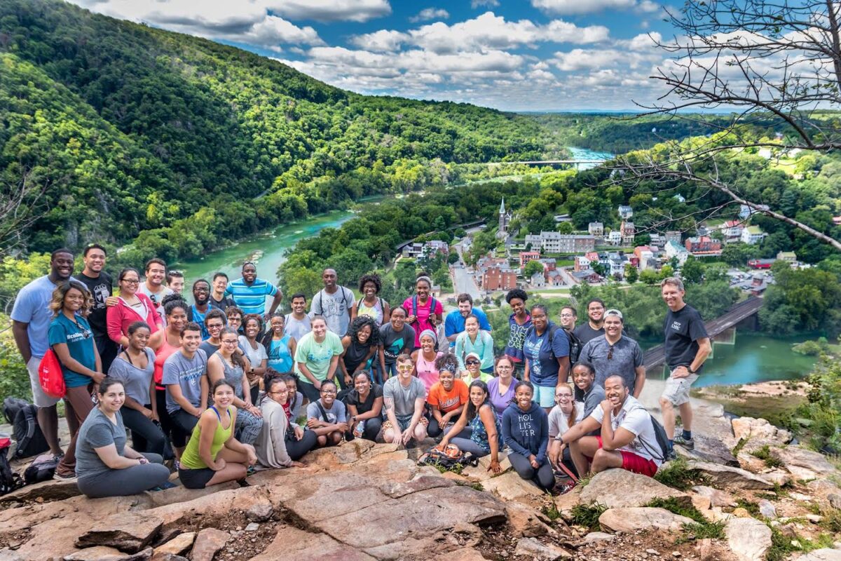 a large group on a rocky outcropping, backed by rolling green hills and a river