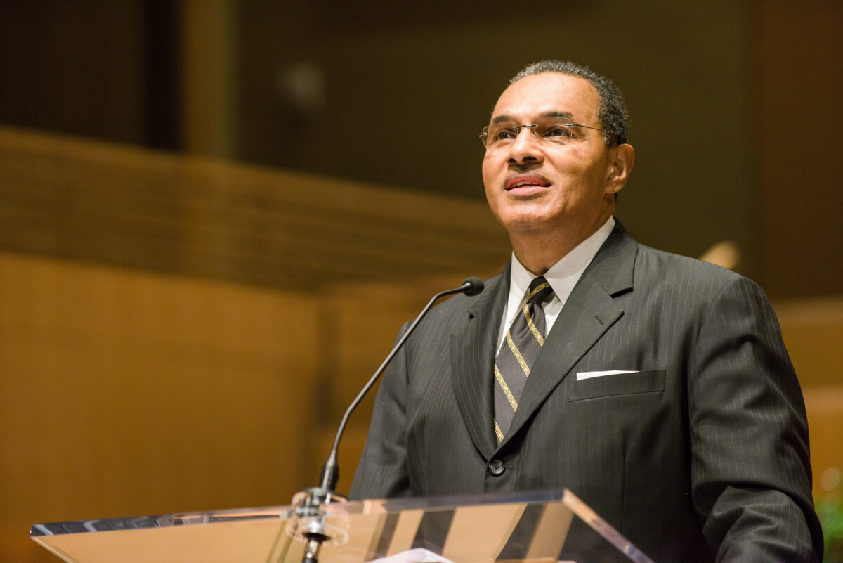 man in suit and black and gold striped tie stands behind a podium with positive thoughtful expression