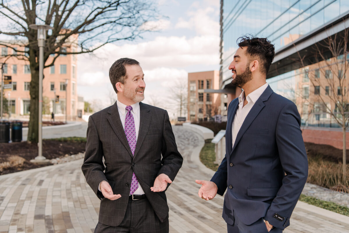 Two adults in suits face each other while standing outside in between buildings.