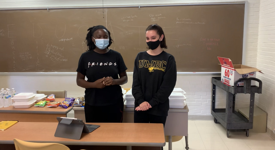 Two young women, one wearing a Friends tv show shirt and another wearing a UMBC Retrievers shirt, standing in front of a snack table.