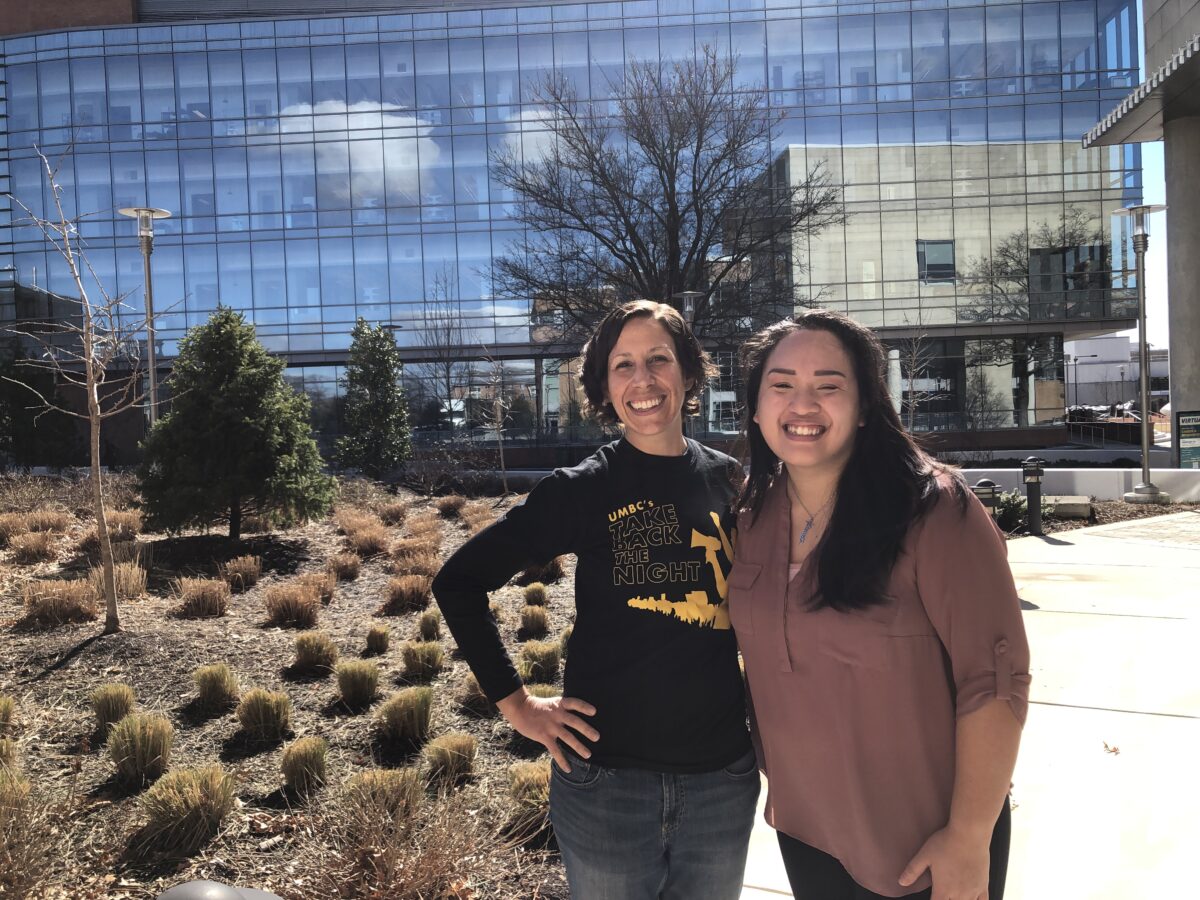 Two women smiling outside in front of glass-paneled building.