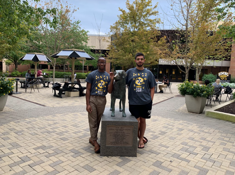 Two adults wearing black t-shirts, stand with a bronze sculpture of a dog at UMBC.