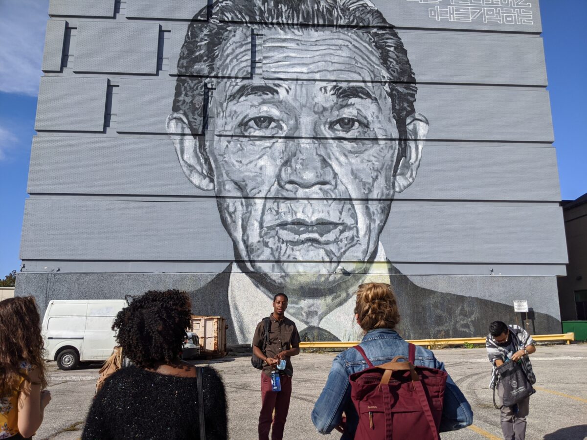 A person facing a group stands in front of a large grey and black mural of a headshot of an adult