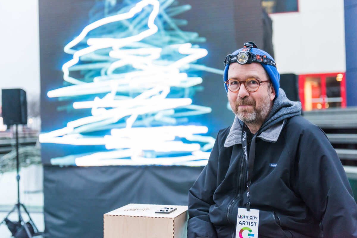 Tim Nohe, wearing a blue hat and coat, and a headlamp, poses in front of artwork that resembles vertical zigzag lines.