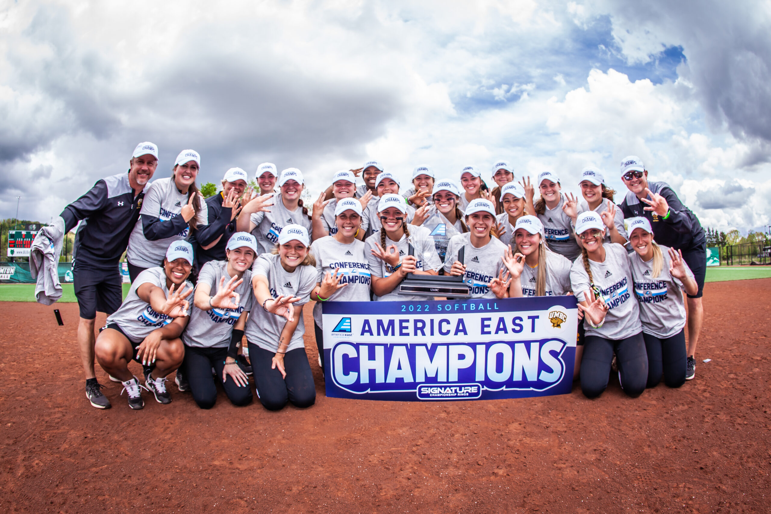 Softball players and coaches smile around "America East Champions" sign, holding 3 fingers up.