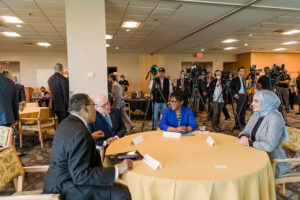 Four people in professional clothes sit at a table, with journalists and cameras in the background, facing them