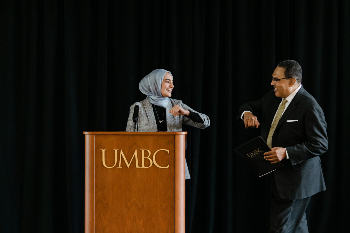 Young woman in sky blue hijab and sport coat stands behind a podium labeled "UMBC," greeting man in suit