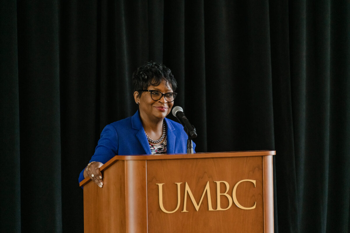 Woman in blue blazer stands behind a podium labeled "UMBC," smiling