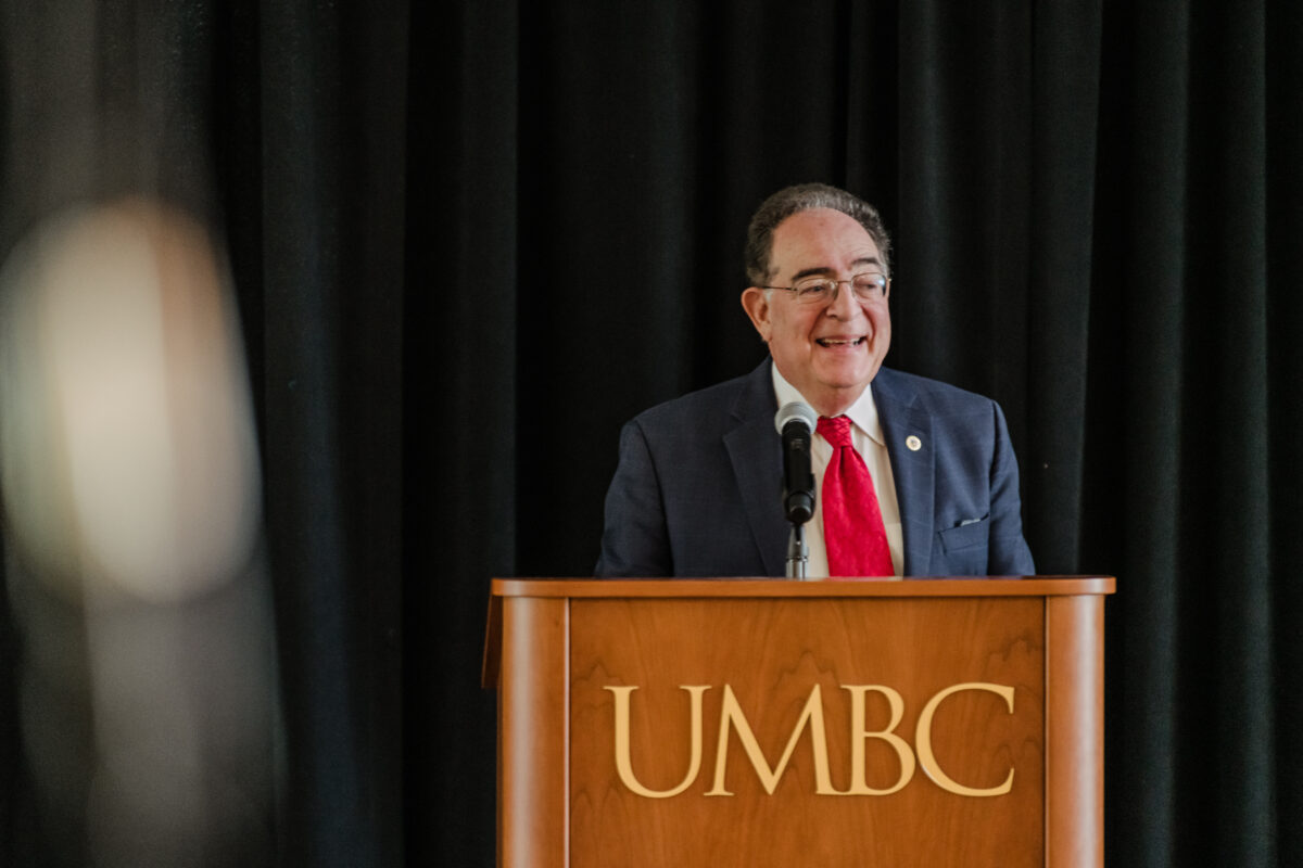 Man in suit and red tie stands behind a podium reading, "UMBC."