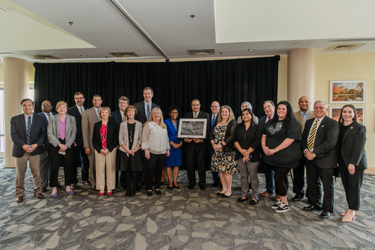 21 adults wearing a mix of professional and everyday clothing stand, with one holding a framed photo.