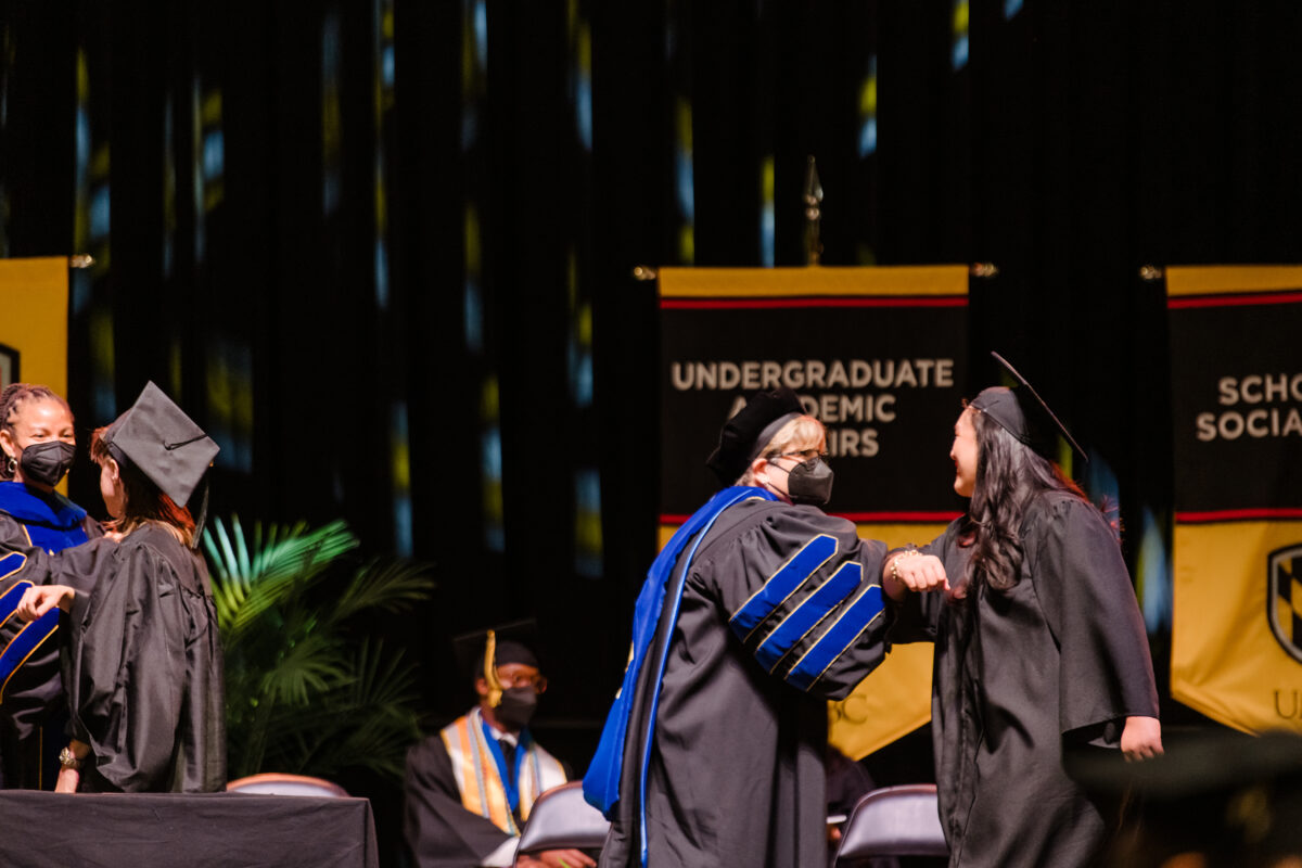 Woman crossing graduation stage in regalia stops to elbow bump a man