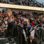 Man wearing face mask walking in commencement regalia waving at audience