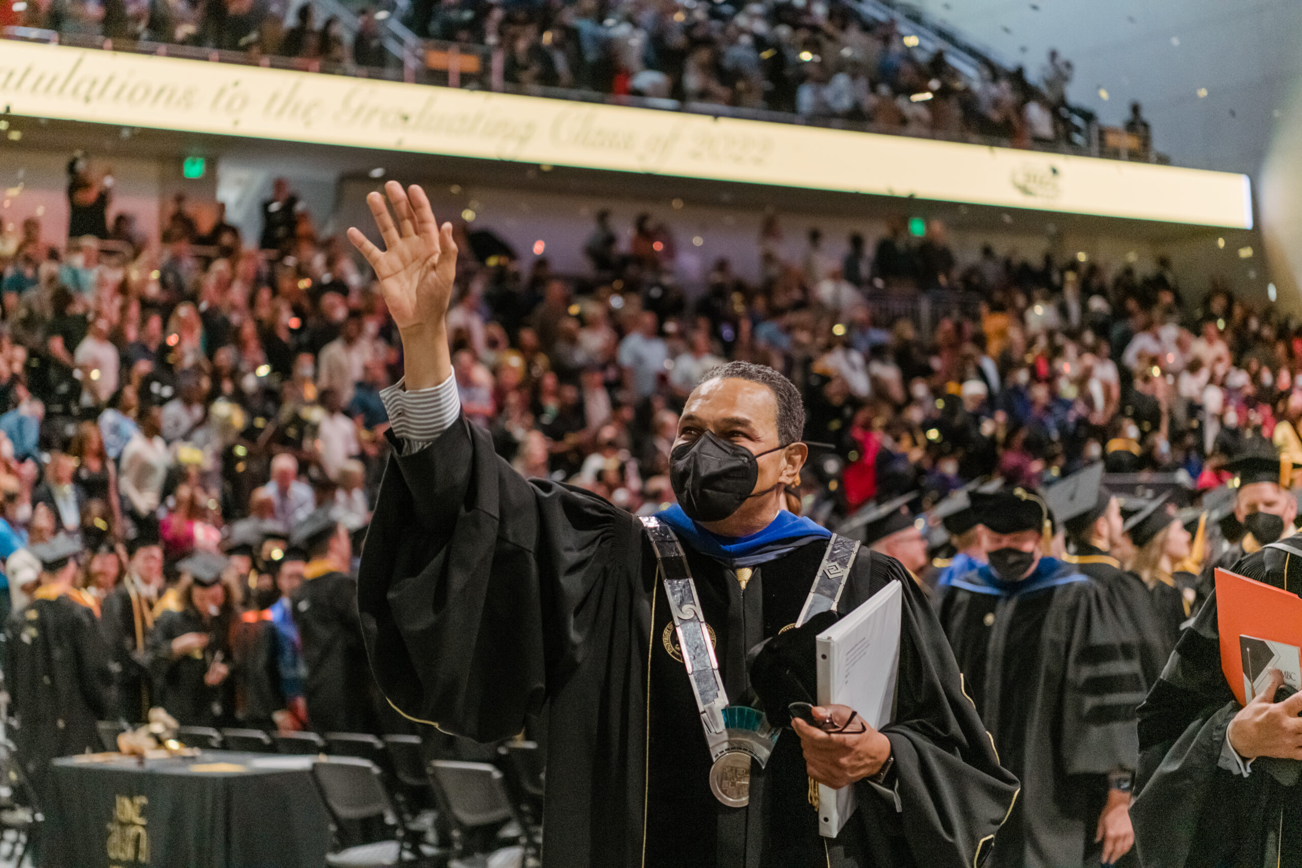 Man wearing face mask walking in commencement regalia waving at audience
