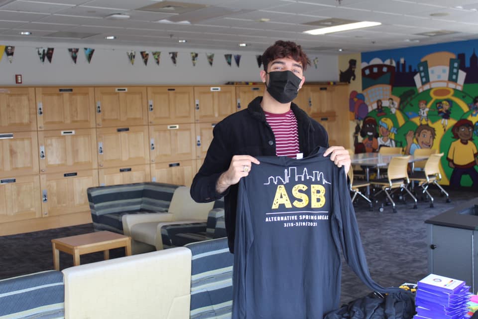A masked adult wearing a purple shirt with white straps and a black cardigan holds up a black t-shirt with the letters ASB written on it in gold.