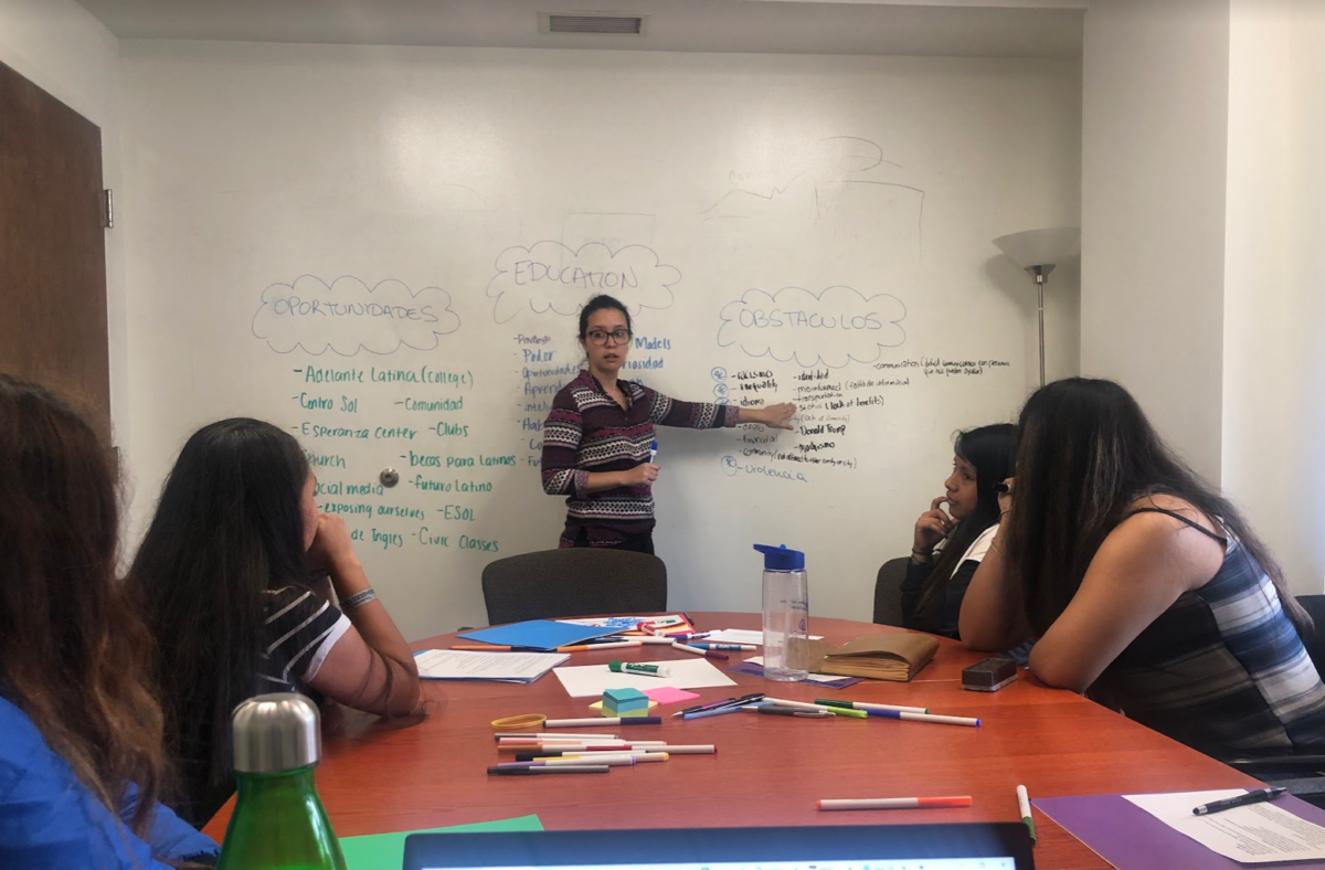 A group of students sit around a red table with books and papers face a person in front of the table with a white board behind them. Immigrant, UMBC, teenager.