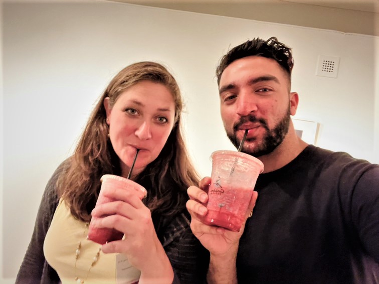Two people stand next to each other sipping a pink drink from a plastic cup.