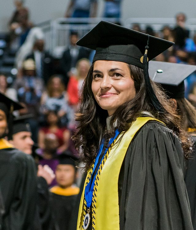 A young woman in her 30s wears a cap and gown, amongst other UMBC graduates.