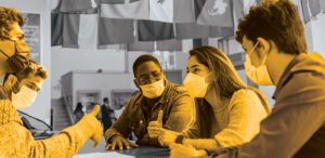 In gold overlay, students sit around a table in the commons talking.