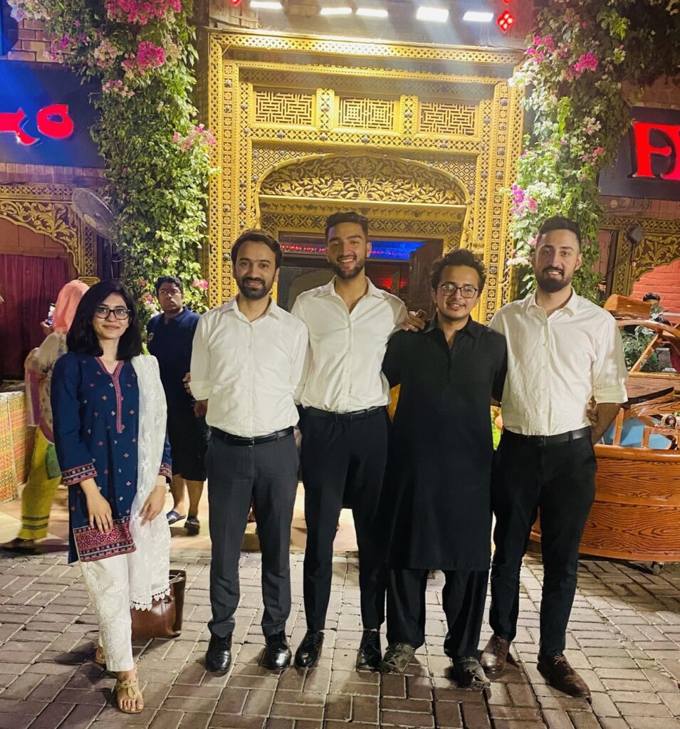 A group of five adults stand next to each other in front of a decorated bright yellow arch.