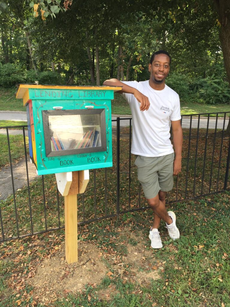 A person wearing shorts and a t-shirt leans on a green wooden box held up by a post. Baltimore.