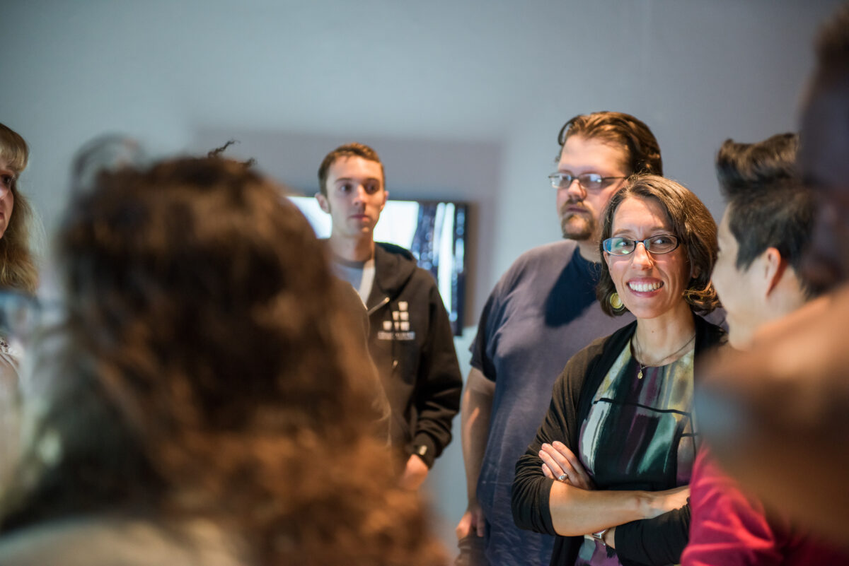 Fulbright award recipient Corrie Parks, wearing glasses and a shirt with water color print, stands with a group of other people in an exhibit space