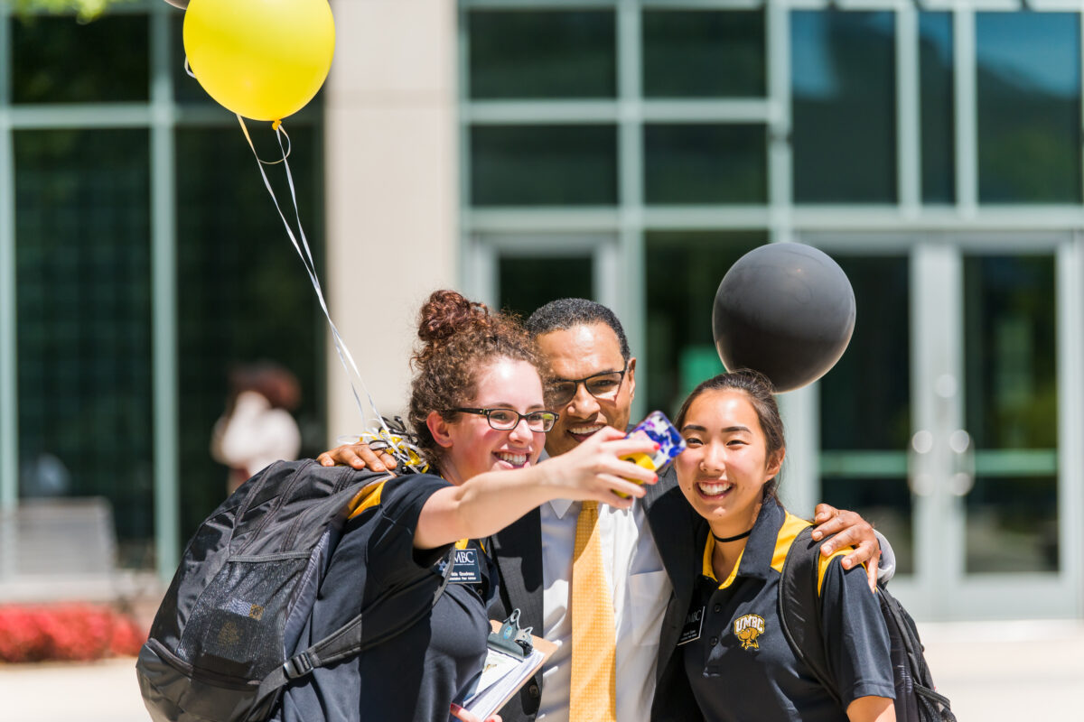 man in black suit with golden yellow tie smiles for a selfie with two students, both wearing UMBC shirts and carrying backpacks and balloons.