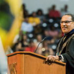 man in dark graduation robe stands behind a podium with positive expression