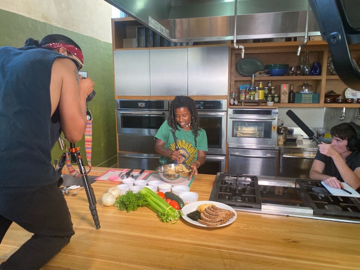 One person with a camera stands on top of a table filming a person cooking while another person holds a microphone. Photo by López. New Orleans.