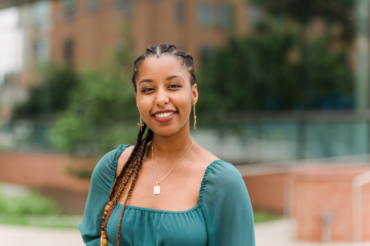 A person with long braided hair wears a green blouse, long golden earrings and necklace, stands in front of trees.