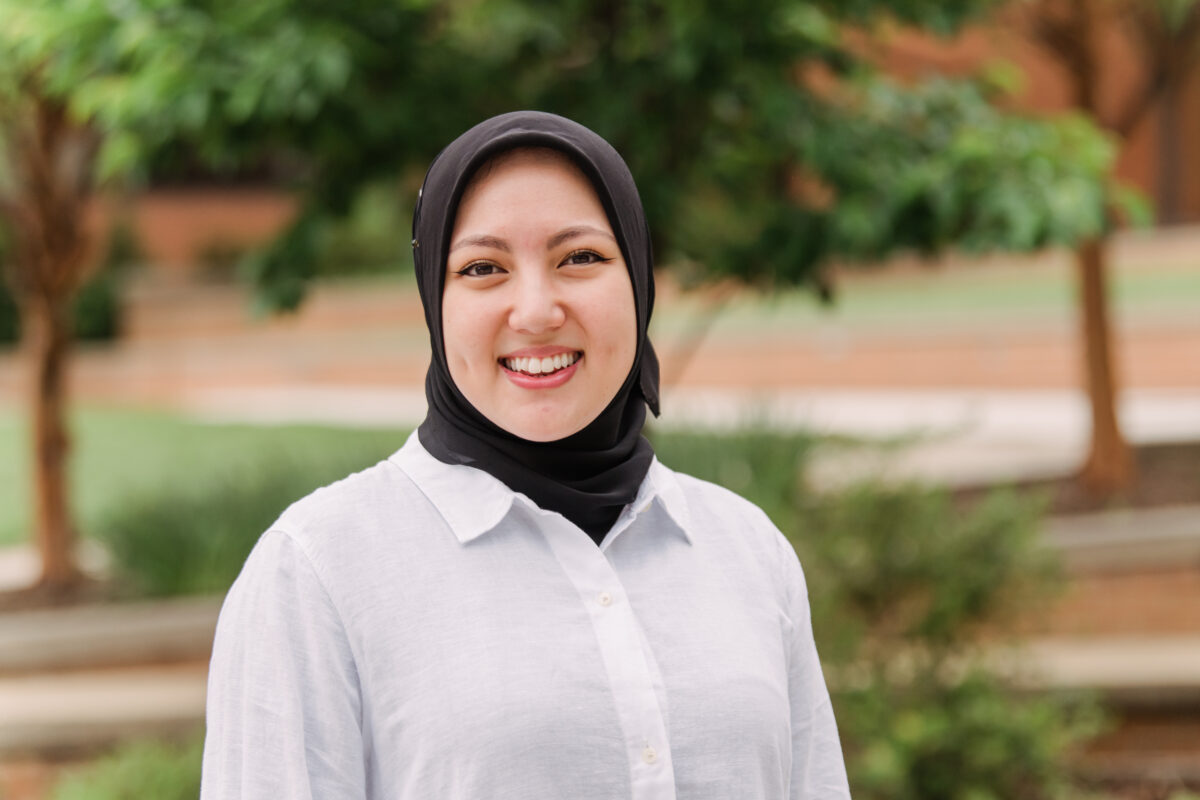 A woman wearing a black hijab and white long sleeve blouse stands outside with trees in the background.