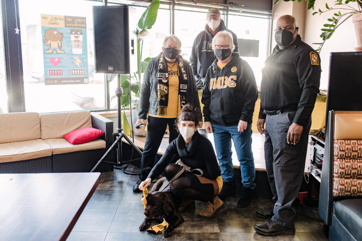 A group of 5 people stand in a coffee shop around a chocolate lab dog laying on the ground