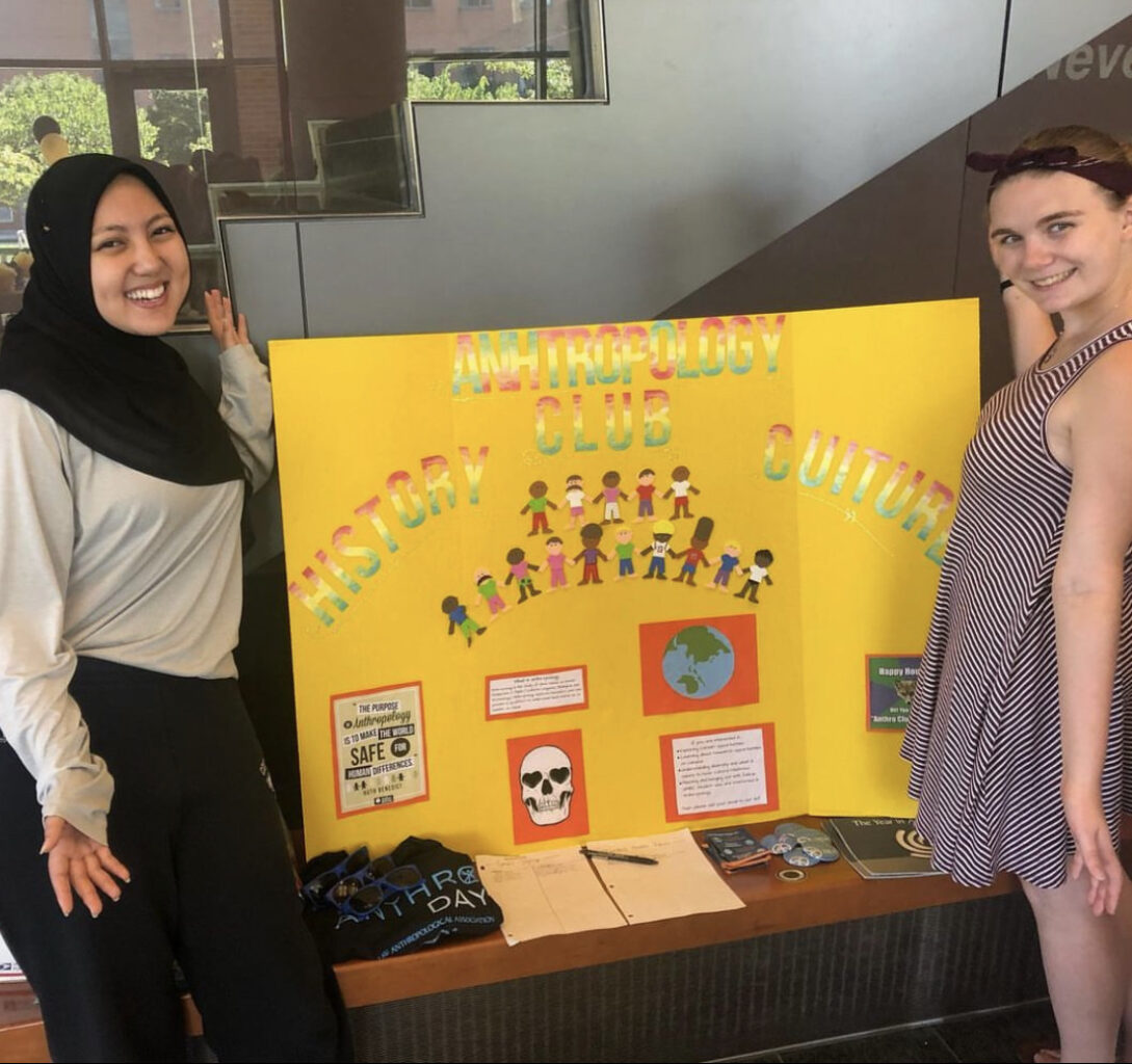 Two adults, one wearing a black hijab and a beige long sleeve shirt and the other wearing a headband with a maroon and white striped tank top stand on each side of a yellow poster with rainbow lettering.