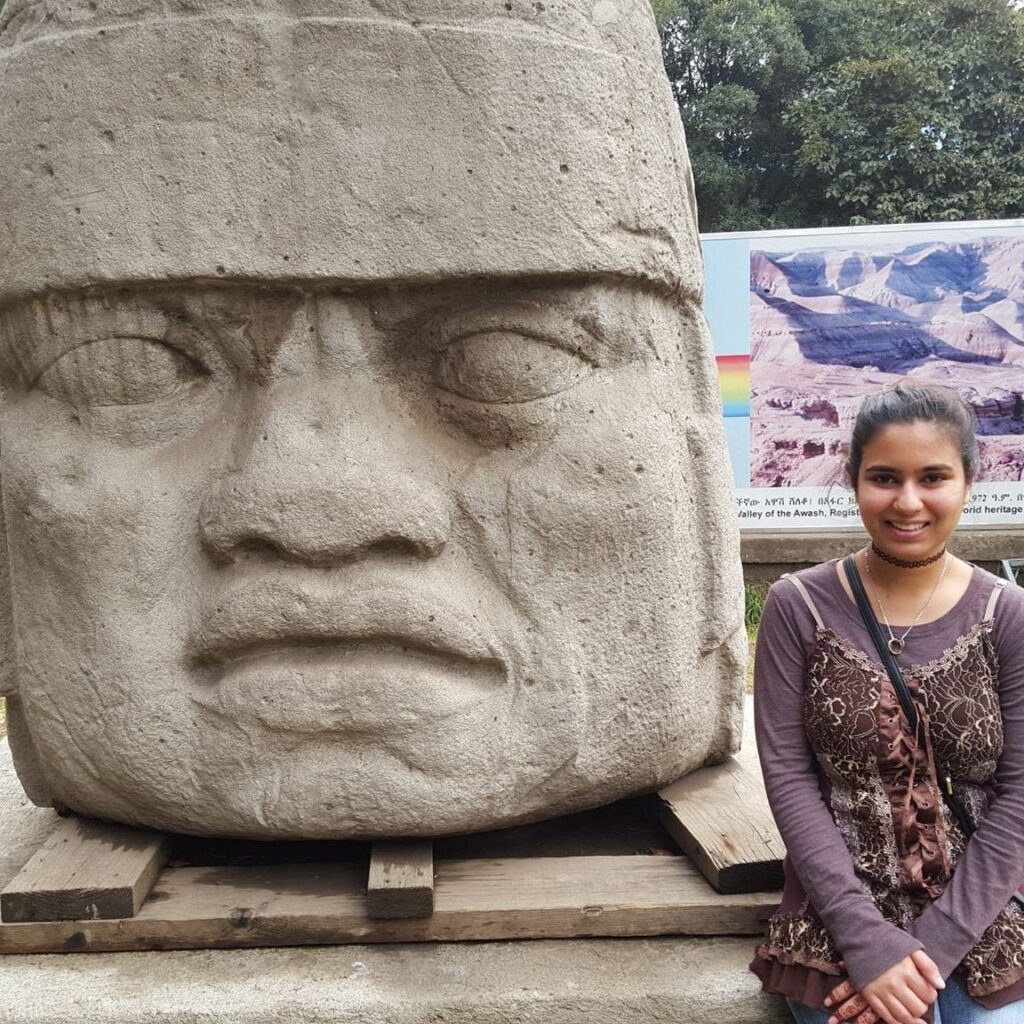 An adult wearing a purple long sleeve shirt and a black purse sits next to a sculpture of an immense stone head.