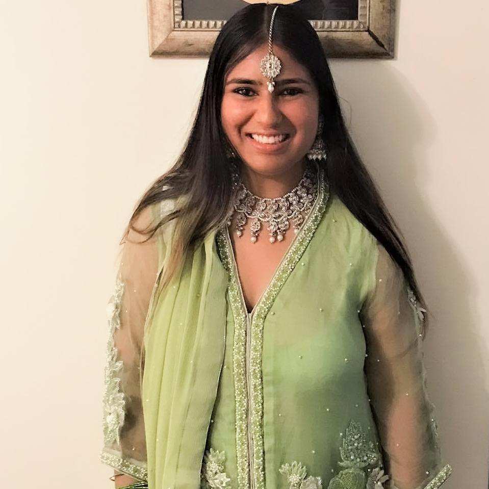 A person with long black hair wears a light green embroidered dress with a beaded necklace and a beaded pendant hanging on their forehead.