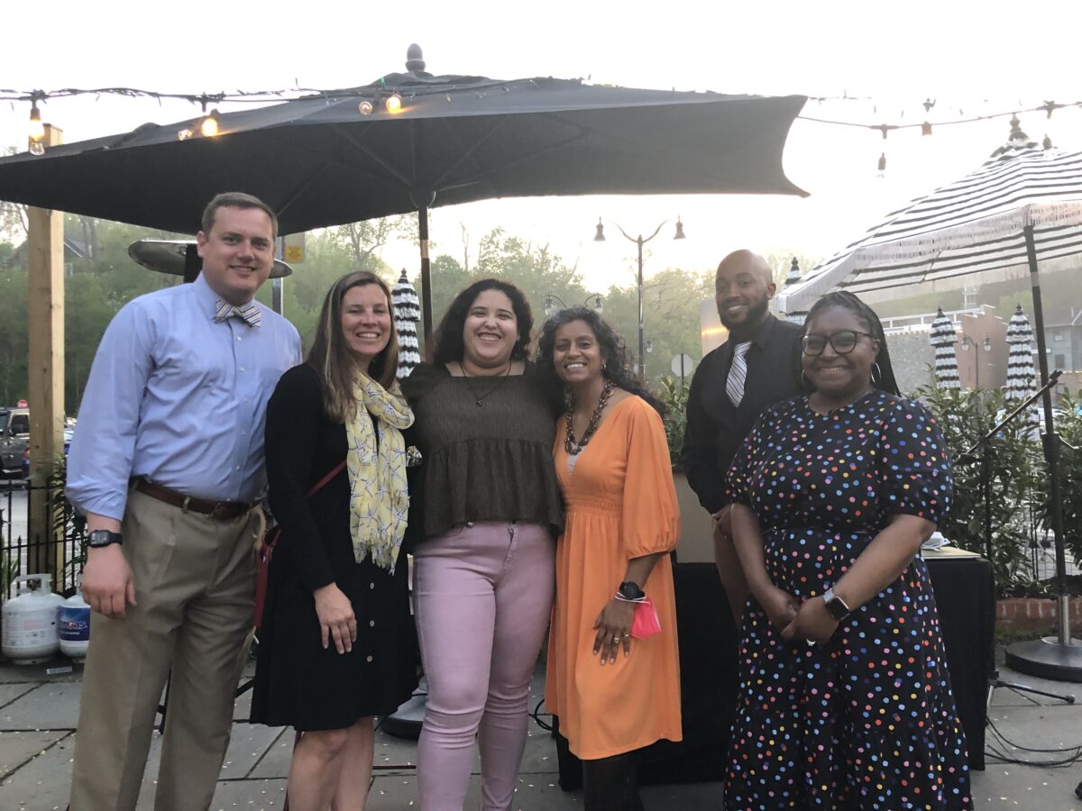 Six adults wearing dressy clothing stand close together under a large black umbrella at a restaurant.