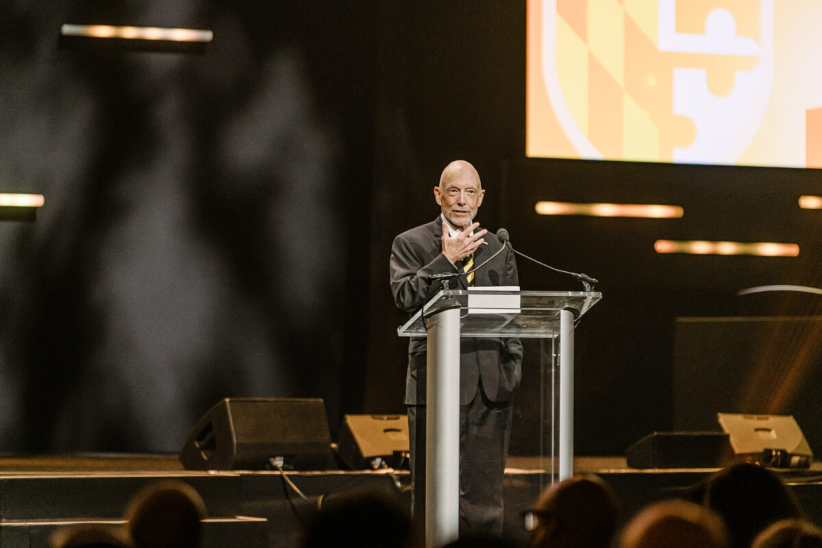 Man wearing a black suit and black and gold striped tie stands, speaking, at a clear podium