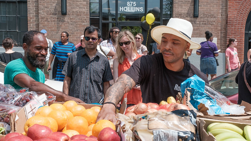 A person wearing a white hat reaches for oranges in a food cart with people in the background.