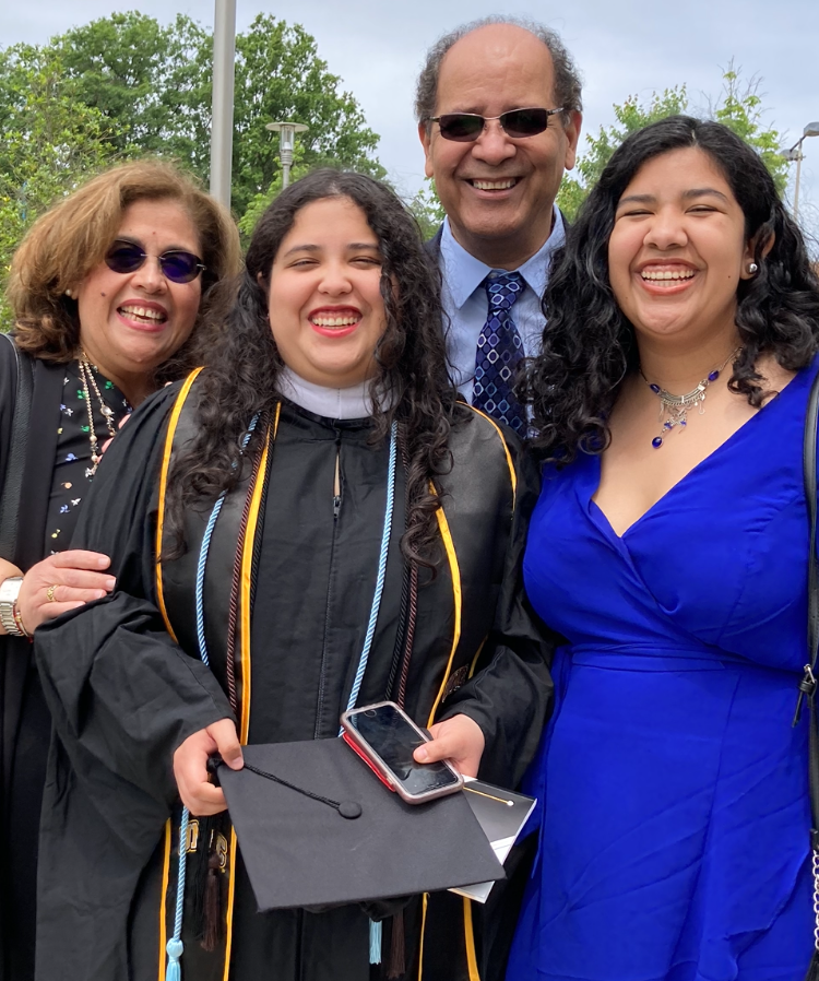 One adult wearing a black graduation cap and gown stands next to a person with a cobalt blue dress with two other adults behind them.