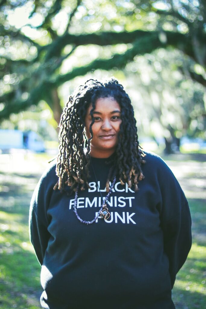 A woman with long black dreadlocks wearing a black sweatshirt with white letters stands outside with grass in the background. Photo by Fernando López