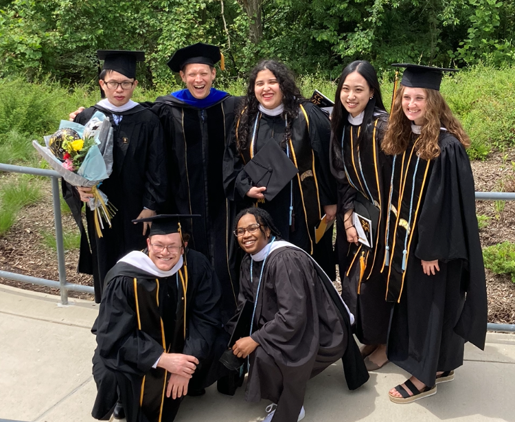 Seven adults wearing black graduation caps and gowns huddle while standing on a path with trees in the background.