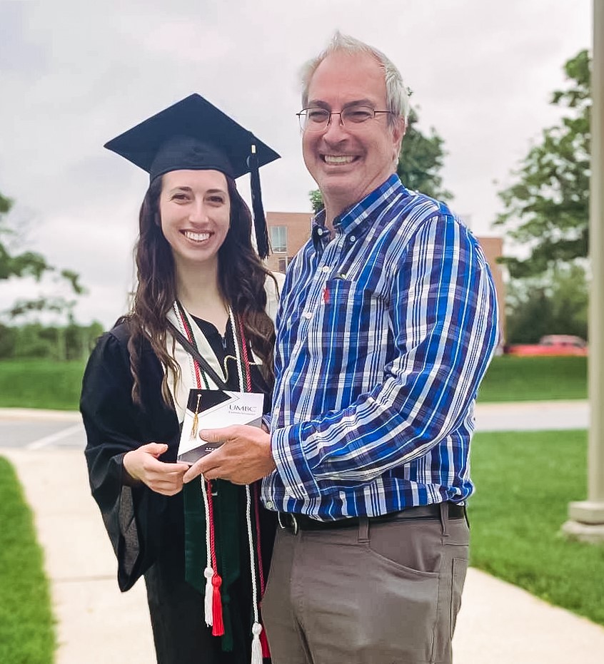 A person wearing a black graduation cap and gown stands outside next to a person wearing a blue plaid dress shirt and grey pants. Both are holding a plaque.