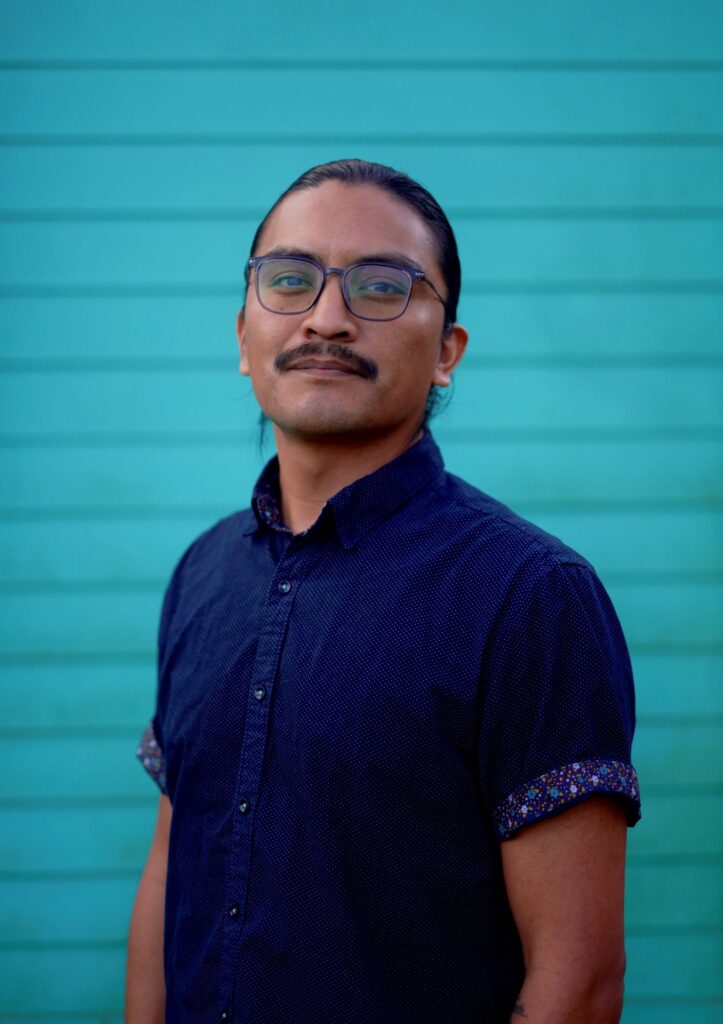 A person with black hair pulled back wearing a cobalt blue dress shirt stands in front of an aqua wall. Photo by Fernando López.