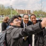 Two adults in professional attire pose for a selfie with a young adult outdoors, smiling. All wear light jackets.