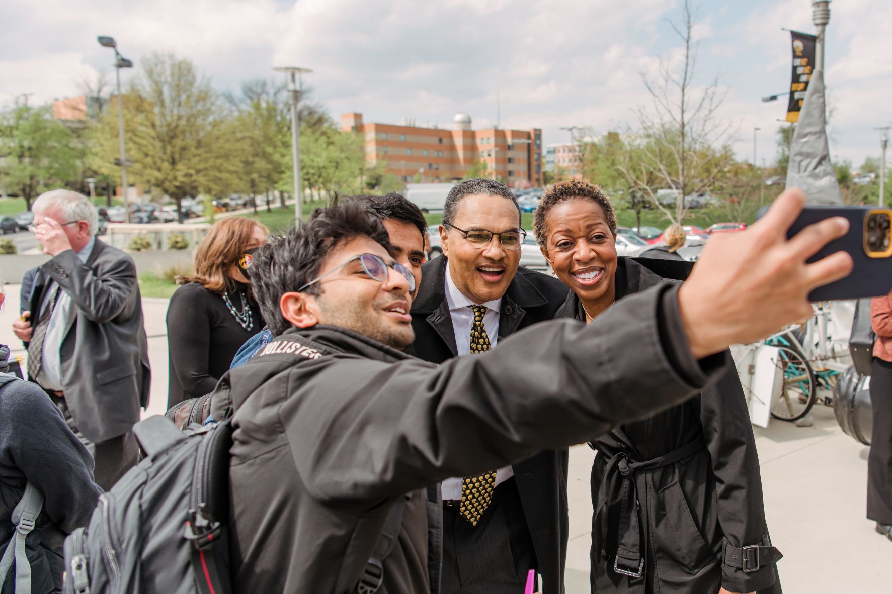 Two adults in professional attire pose for a selfie with a young adult outdoors, smiling. All wear light jackets.