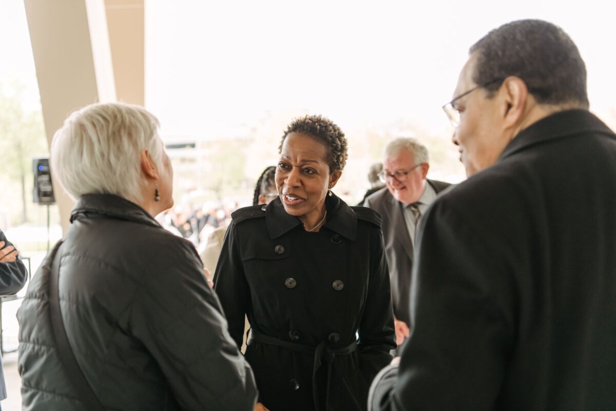 Woman in black coat shakes hands with another woman while two men look on.