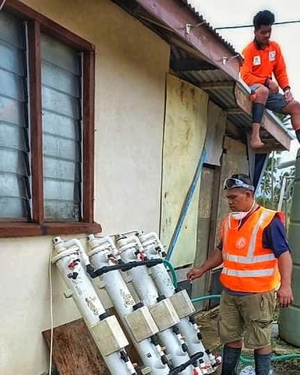 A person wearing a bright orange and white vest and shorts stands next to white pipes that are leaning on a house.