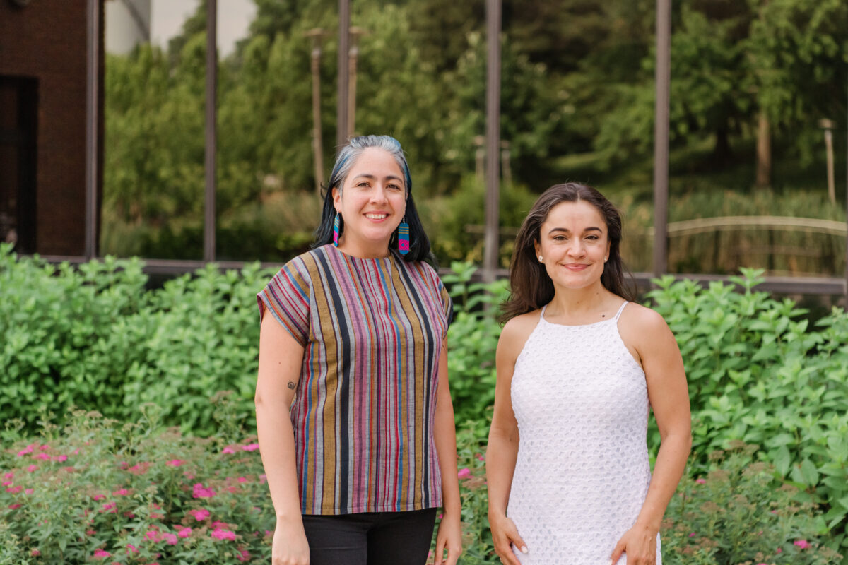 Two people standing side by side, one is wearing a stripped multicolored short sleeve shirt and the other is wearing a white dress, they are standing in front of a wall of windows and shrubbery.