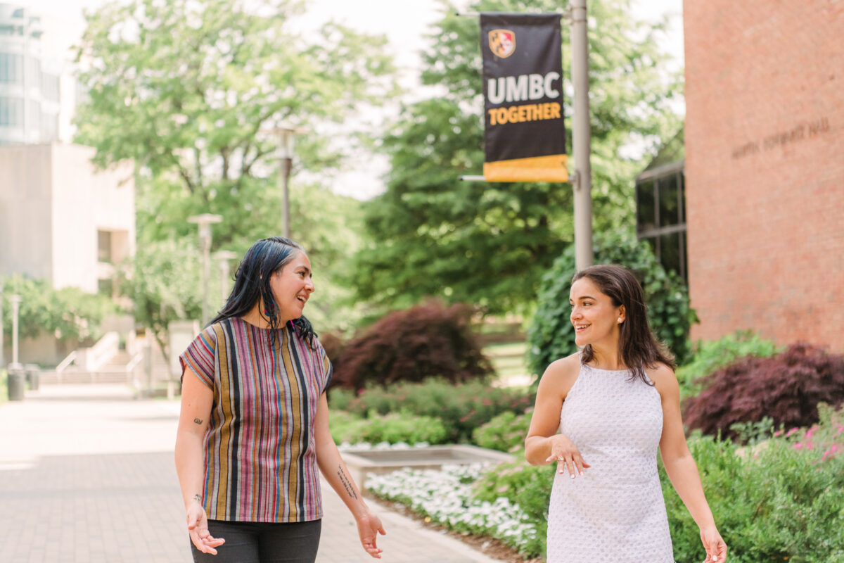 Two people walk side by side down a brick pathway with landscaped shrubbery behind them, one is wearing a multicolored striped shirt and the other a white dress. UMBC Mellon